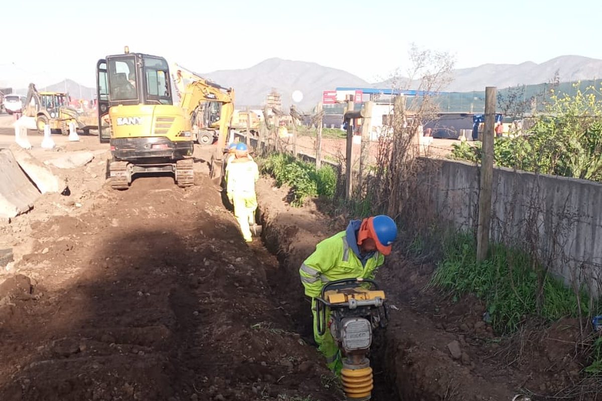 Excavación de zanja para canalización con retroexcavadora y cuadrilla de MCF compactando el fondo.